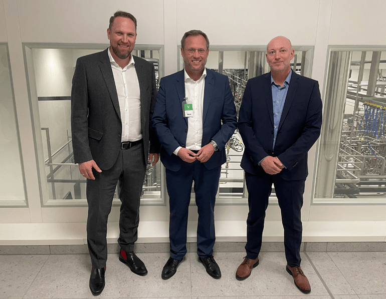 Martin Ansbacher steht zusammen mit Andreas Burkhardt und Stefan Fügenschuh vor einer großen Werkshalle, in der sich technische Anlagen befinden.