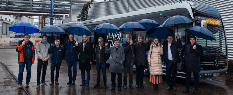 Eine Gruppe von Menschen mit nlauen Regenschirmen vor einem Wasserstoffbus