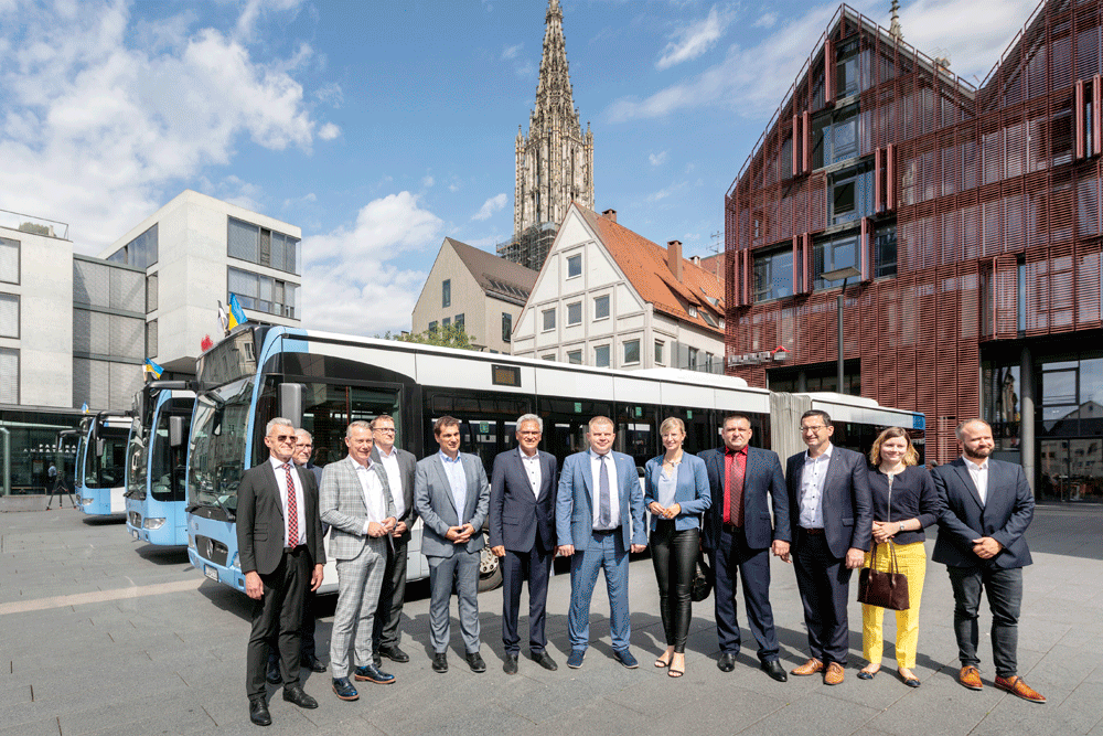 Drei Busse parken auf dem Hans-und-Sophie-Scholl-Platz, davor haben sich zwölf Personen in förmlicher Kleidung mit Anzug und Jacket zum Foto aufgestellt.