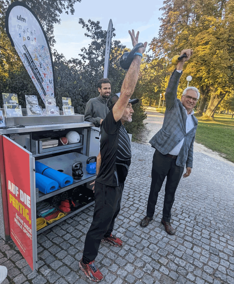 Oberbürgermeister Gunter Czisch und ein Mann in einem schwarzen Trainingsanzug stemmen eine Hantel in die Höhe