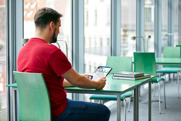 Mann mit rotem T-Shirt sitzt an einem grünen Tisch. Er hat ein Tablet in der Hand auf dem er die Onleihe der Stadtbibliothek Ulm bedient.