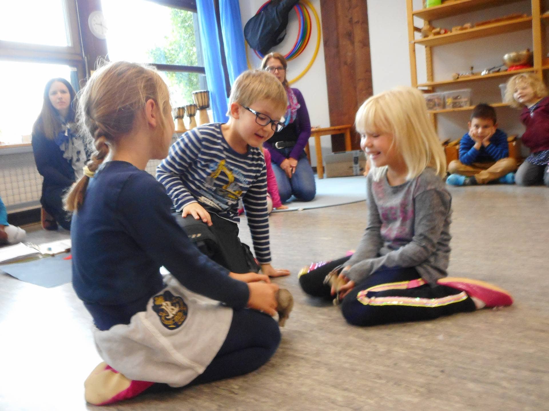 Kindergartenkinder sitzen im Kreis beim Rollenspiel, eine Erzieherin beobachtet im Hintergrund