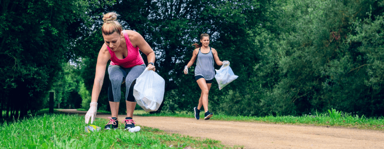 Zwei Frauen mit Müllbeuteln joggen im Park, eine von ihnen hebt gerade eine leere Verpackung auf.