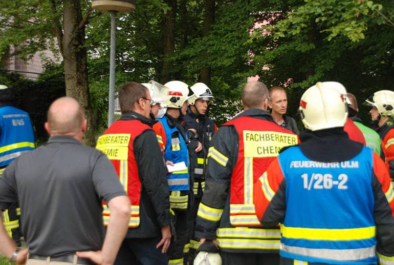 Lagebesprechung der Einsatzleitung der Feuerwehr Ulm bei einem Gefahrguteinsatz. Die Funktionsträger wie der Einsatzleiter, der Zugführer, mehrere Gruppenführer und die Fachberater Chemie sind mit bunten Warnwesten gekennzeichnet.