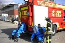 Bei einem Chemieeinsatz sitzen zwei Angehörige der Feuerwehr Ulm in blauen Chemikalien-Vollschutzanzügen auf den Trittbrettern des Großtanklöschfahrzeuges zum Einsatz bereit.