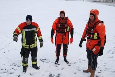 Zwei Eisretter der Feuerwehr Ulm in ihren orangefarbenen Offshore-Trockenanzügen mit Rettungsweste stehen bei einer Rettungsübung auf der beschneiten Eisfläche des Ausees. Der Gruppenfüher weist in die Übung ein.