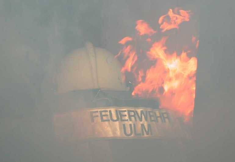 Ulmer Feuerwehrmann in einem brennenden Raum; von hinten ist das Rückenschild mit Feuerwehr Ulm, der Helm und viele Flammen zu sehen.