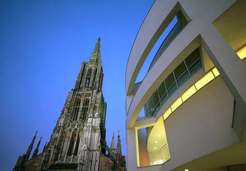Das bogenförmige Außengeländer des Stadthauses erhebt sich zusammen mit dem Turm des Ulmer Münsters in der Dämmerung.