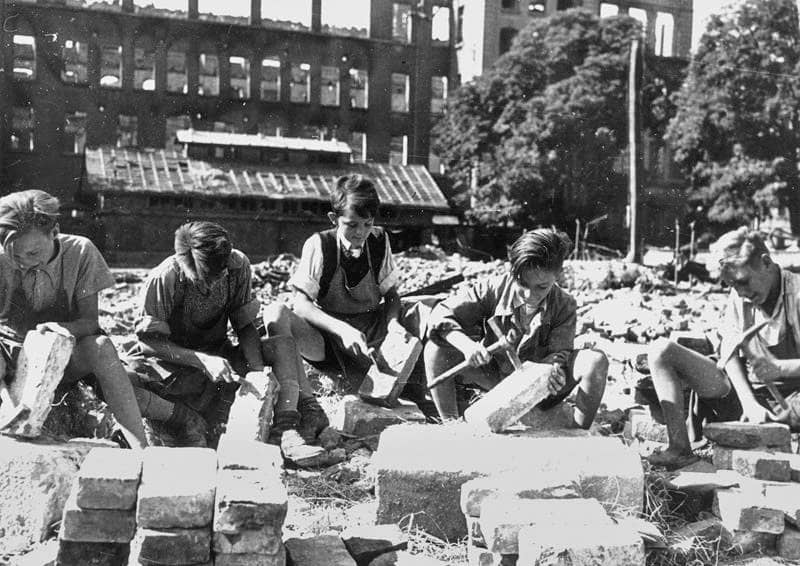 Fünf Jungen zerkleinern Steinbrocken auf dem Platz vor einem zerbombten Haus.