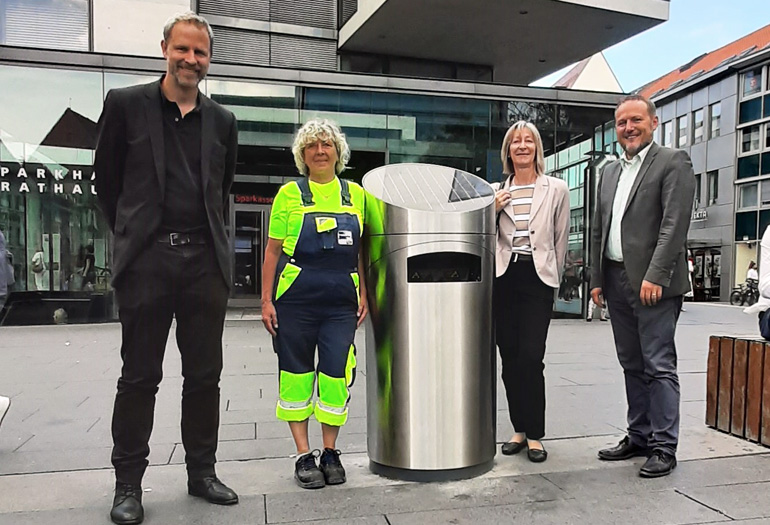 Vier Personen stehen neben dem neuen Mülleimer auf dem Hans-und-Sophie-Scholl-Platz.