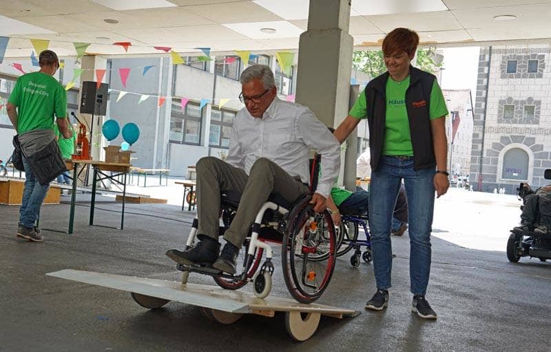 Gunter Czisch fährt in einem Rollstuhl über eine Rampe und wird dabei von einer Caritas-Mitarbeiterin gestützt.