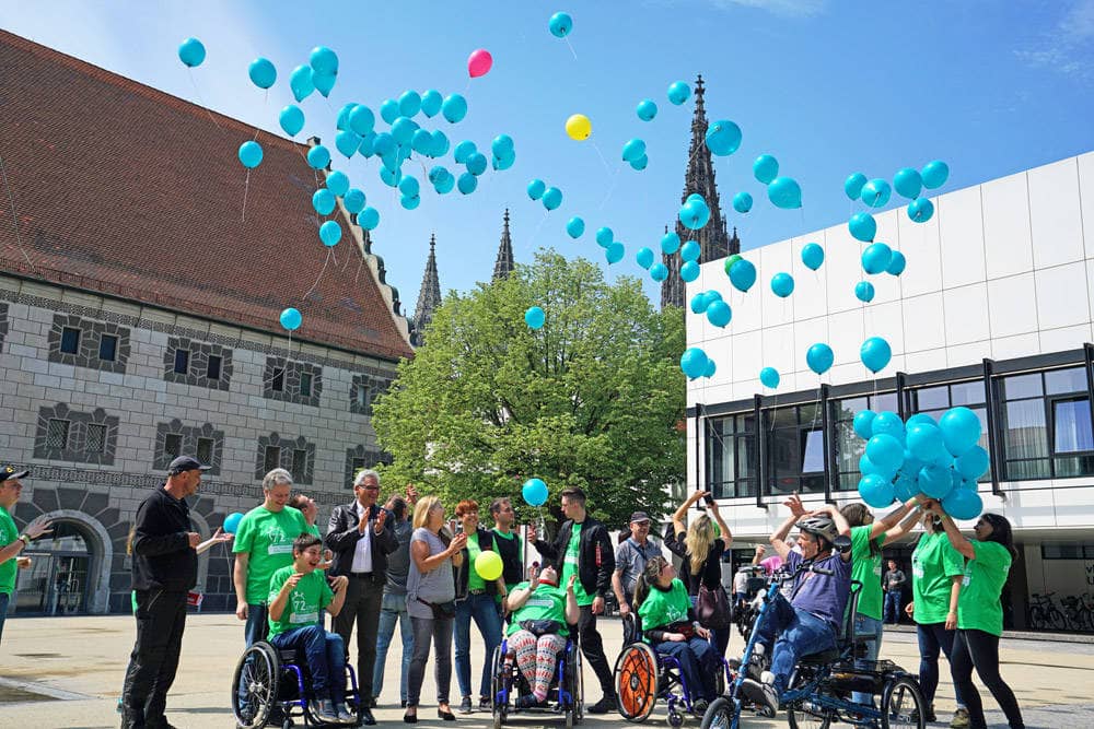 Eine Gruppe von rund 20 Personen, davon einige im Rollstuhl, lassen auf dem Kornhhausplatz Luftballons in den Himmel steigen. Im Hintergrund erhebt sich das Ulmer Münster.