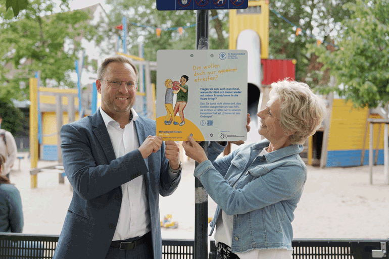 Oberbürgermeister Martin Ansbacher und eine blonde Frau mit Jeansjacke zeigen auf ein Schild vor einem Spielplatz