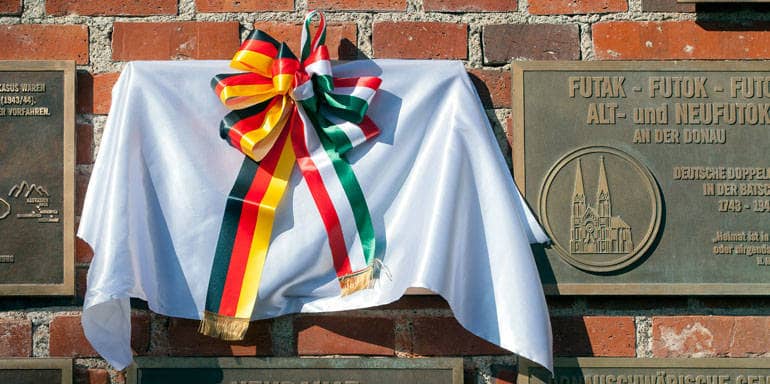 Ein weißes Tuch mit einer Schleife in den Farben der deutschen und der ungarischen Flagge verdeckt eine Tafel an der Stadtmauer.