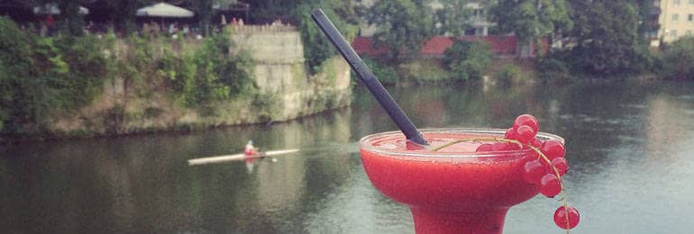 Ein Cocktailglas, das ein rotes Getränk enthält, ist auf der Ulmer Stadtmauer abgestellt. Im Hintergrund fließt die Donau, auf der ein Ruderer entlangfährt.