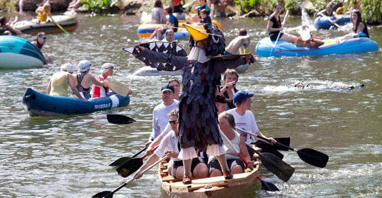 Auf einem kleinen Boot, in dem rund zehn Menschen mit Paddeln sitzen, steht eine Person in einem den ganzen Körper verhüllenden Spatzen-Kostüm.