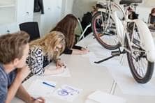 Mehrere Studieren zeichnen Entwürfe eines Fahrrades an einem Tisch, in dessen Mitte ein Fahrrad aufgestellt wurde.