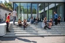 Auf den Treppenstufen vor dem Hochschulgebäude sitzen Studierende in kleinen Gruppen beieinander und unterhalten sich.