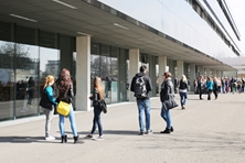 Vor dem Eingangsbereich des Hochschulgebäudes stehen und laufen Studierende in Grupen.