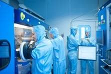 Vier Männer in blauen Schutzanzügen stehen im Labor, zwei von ihnen greifen mit Schutzhandschuhen in einen durch Glas abgedichteten Bereich.