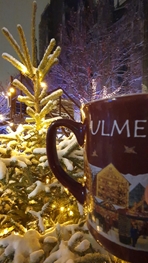 Eine Person hält eine Tasse mit Glühwein in der Hand. Im Hintergrund ein Weihnachtsbaum und das Ulmer Münster.