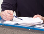 Zwei Hände einer Person halten einen Stapel Papiere und deuten mit einem Stift auf eine Stelle des obersten Blattes.