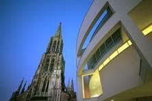 Das bogenförmige Außengeländer des Stadthauses erhebt sich zusammen mit dem Turm des Ulmer Münsters in der Dämmerung.