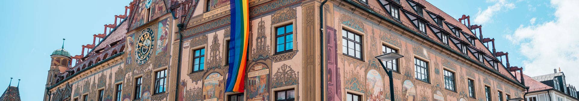 Ausschnitt des historischen Gebäudes des Ulmer Rathauses mit seiner astronomischen Uhr und einer Regenbogenfahne.