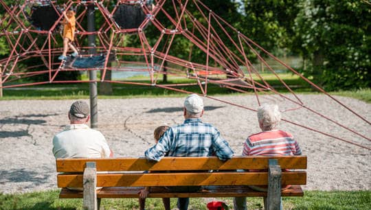 Drei Senioren sitzen auf einer Parkpank vor einem Spielplatz.