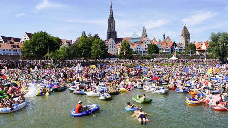 Viele feiernde Menschen lassen sich in Schlauchbooten die Donau hinabtreiben, während am Ufer hunderte von Zuschauern dem Spektakel zusehen.