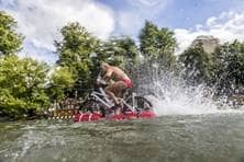 Ein Nabada-Teilnehmer bewegt sich mit einem Fahrrad, das auf einem Schlauchboot befestigt ist, über das Wasser.