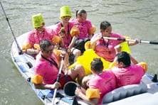 Eine Gruppe von Menschen mit bunten Schwimmflügeln und Eimern auf dem Kopf paddelt in einem Schlauchtboot.