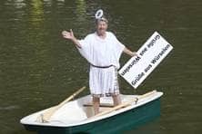 Ein älterer Herr mit Engelskostüm steht auf einem kleinen Boot und grüßt mit einem Schild, auf dem steht: "Grüße aus Würselen".