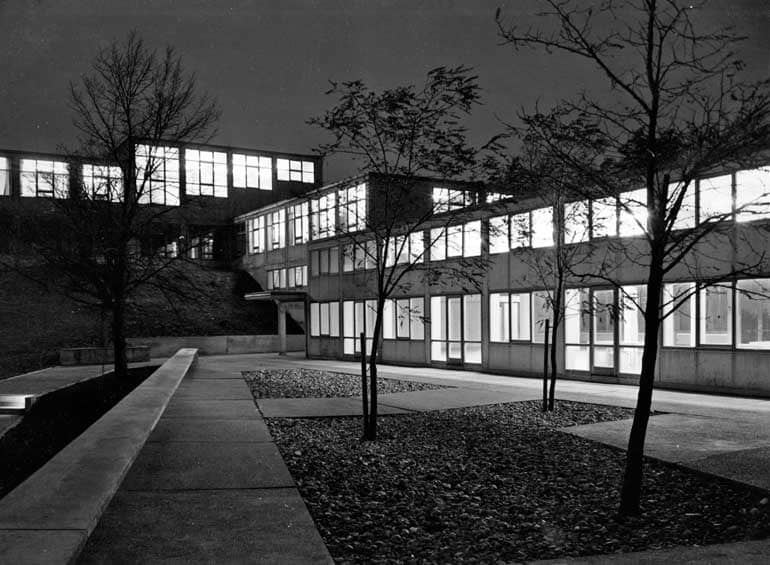 Schwarz-weißes-Foto von rechteckigen Gebäuden mit großen Fenstern, aus denen helles Licht in die Dunkelheit strahlt. Der Platz davor ist zurückhaltend in rechtwinklige Rasen- und Gehwegflächen gehalten.