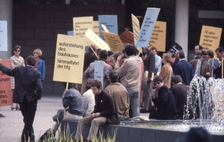 Eine Gruppe von Leuten demonstiert mit Plakaten vor einem Gebäude. Auf einem der Plakate ist zu lesen: "auferstehung des bauhauses - himmelfahrt der hfg".