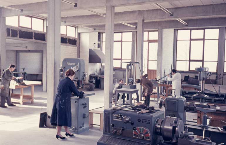 Innenansicht einer hellen Werkshalle mit hohen Fenstern und Betonpfeilern. Auf dem Boden stehen Maschinen und Tische, an denen ein paar Männer und eine Frau technische Arbeiten verrichten.