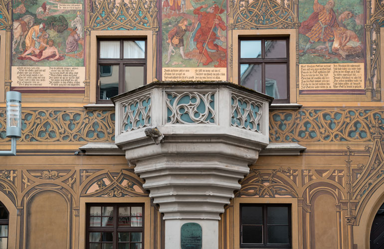 Grauer Balkon im ersten Stock des Rathauses, dessen Geländer mit kunstvoll geschwungenen Steinbögen verziert ist.