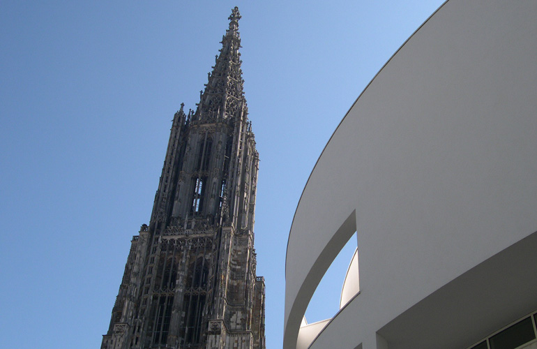 Hauptturm des Ulmer Münsters mit Spitze und Fassade des weißen Stadthauses, die im gezeigten Bereich rundlich ist.