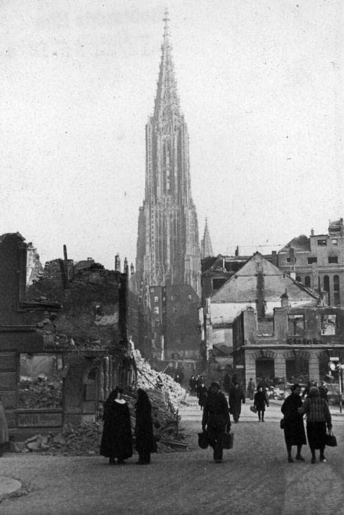 Schwarz-weiß-Foto von Trümmern und eingerissenen Häuserfassaden, im Hintergrund der Hauptturm des Münsters. Auf der Straße sind Menschen mit Taschen unterwegs und sprechen miteinander.