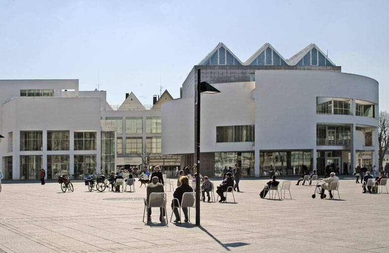 Auf dem Münsterplatz stehen vereinzelt robuste Stühle, auf dem Menschen Platz genommen haben. Am Rande des Platzes erhebt sich das Stadthaus mit seiner runden Fassade und spitzen Dachgiebeln.