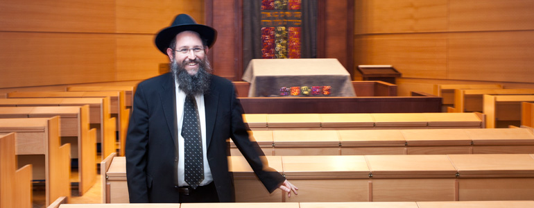 Ein Rabbiner mit langem Bart, schwarzen Hut, Anzug, Brille und ansteckendem Lächeln steht im Innenraum der Synagoge in einer Reihe von Gebetsbenken.
