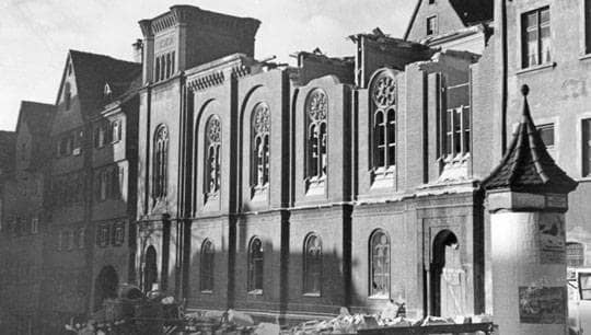 Schwarz-weiß-Foto der alten Synagoge, deren Dach abgerissen worden ist, sodass die Innenwände oben herausragen. Auf der Straße liegen Trümmer.