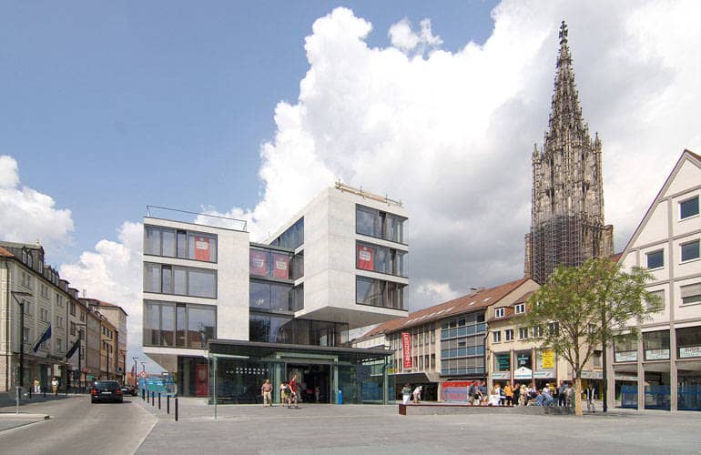 Ein weitläufiger Platz, an dessen Ende ein Gebäude - bestehend aus mehreren Quadern mit hohen Glasfenster - steht. Im Hintergrund erhebt sich der Hauptturm des Ulmer Münsters.