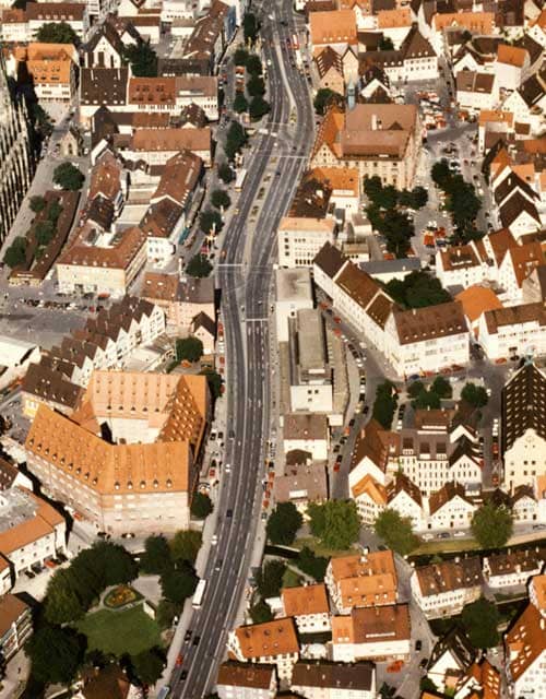 Hochformatige Luftaunfnahme, durch die sich von oben bis unten eine breite Straße zieht. Links und rechts davon sind Dächer der Ulmer Innenstadt zu sehen.
