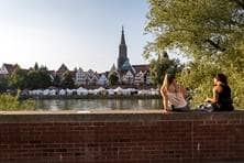 Zwei junge Frauen sitzen auf der Stadtmauer in Neu-Ulm und schauen auf die andere Seite der Donau nach Ulm.