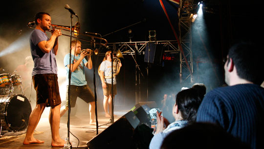 Ein junger Posaunist und zwei weitere Musiker stehen auf der Bühne und heizen dem Publikum ein.